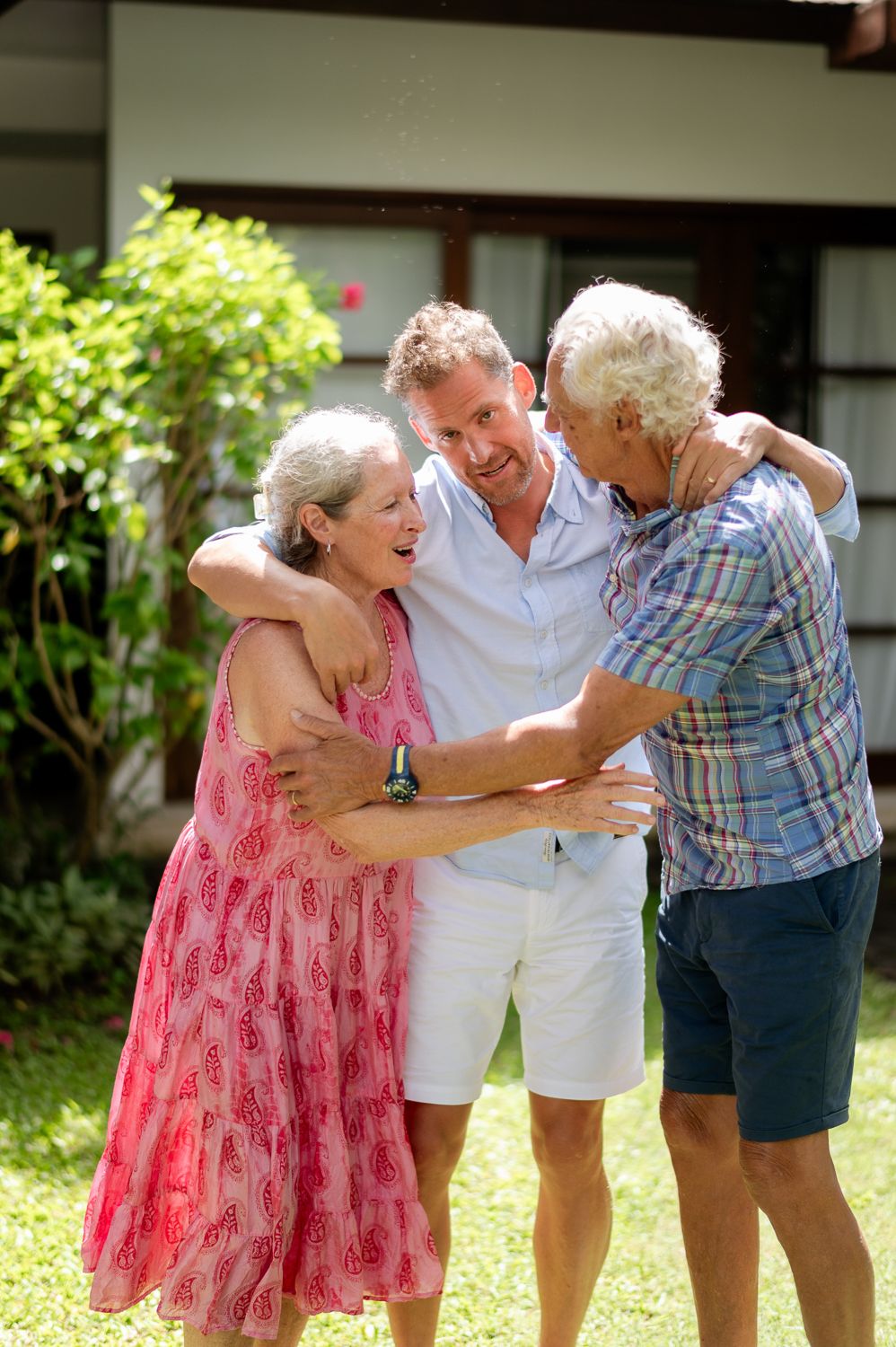 Three seniors share a warm embrace outdoors, smiling joyfully in a sunny garden near a white house.