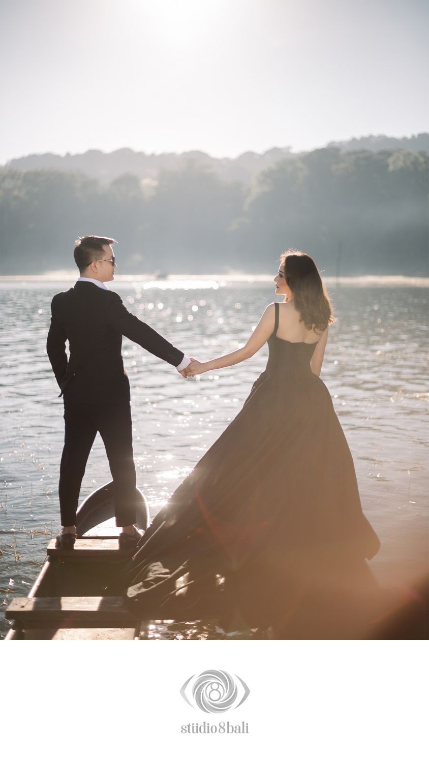 Couple in black attire holding hands on a dock by a sunlit lake.