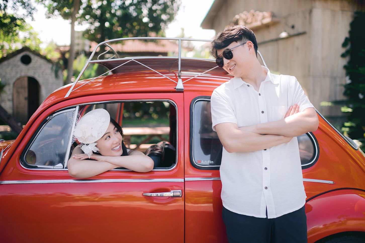 Couple posing with a vintage red Fiat 500, woman leaning out window wearing white hat, man in white shirt with sunglasses.