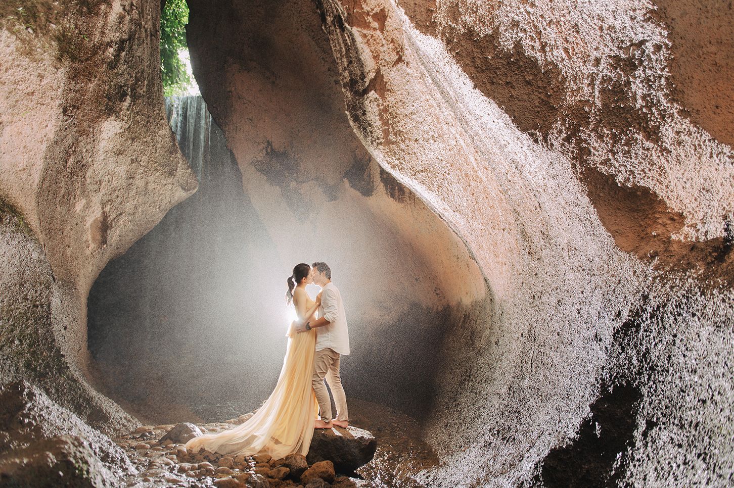 Couple kissing in a sunlit cave with misty waterfall, woman in cream gown, romantic Bali pre-wedding photography.