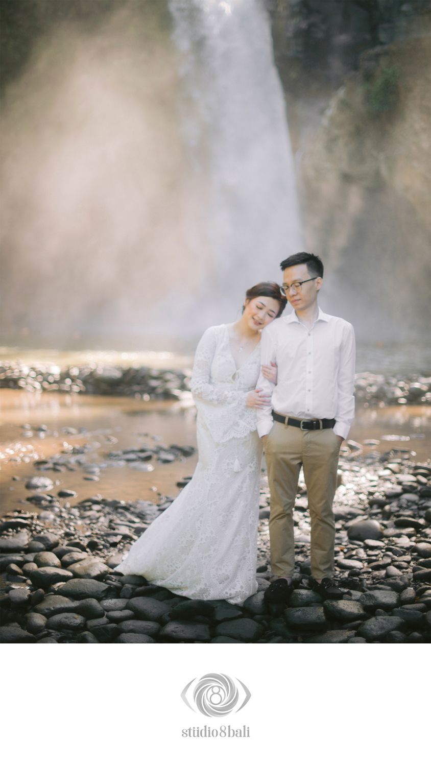 Couple in white wedding attire poses romantically by a misty waterfall on rocky terrain in Bali.
