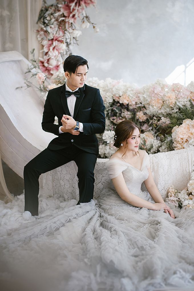 Couple in elegant wedding attire pose among floral decorations, groom in tuxedo and bride in white lace gown.