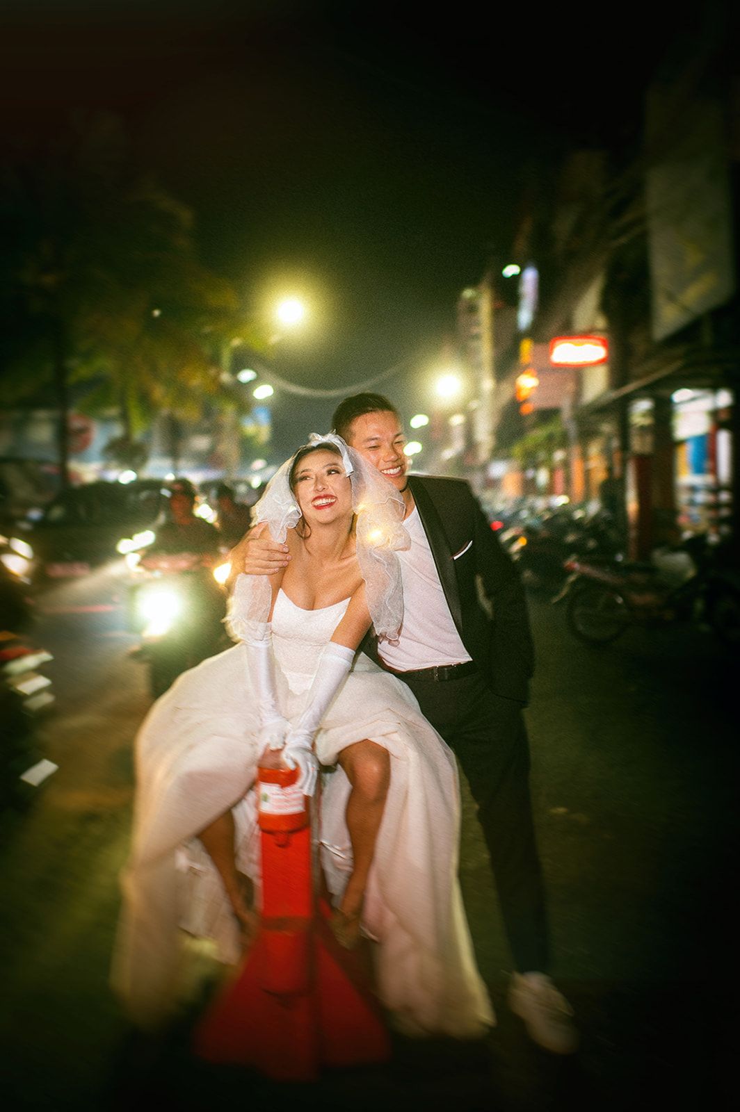 Groom embracing bride seated on a fire hydrant on a lively city street at night.