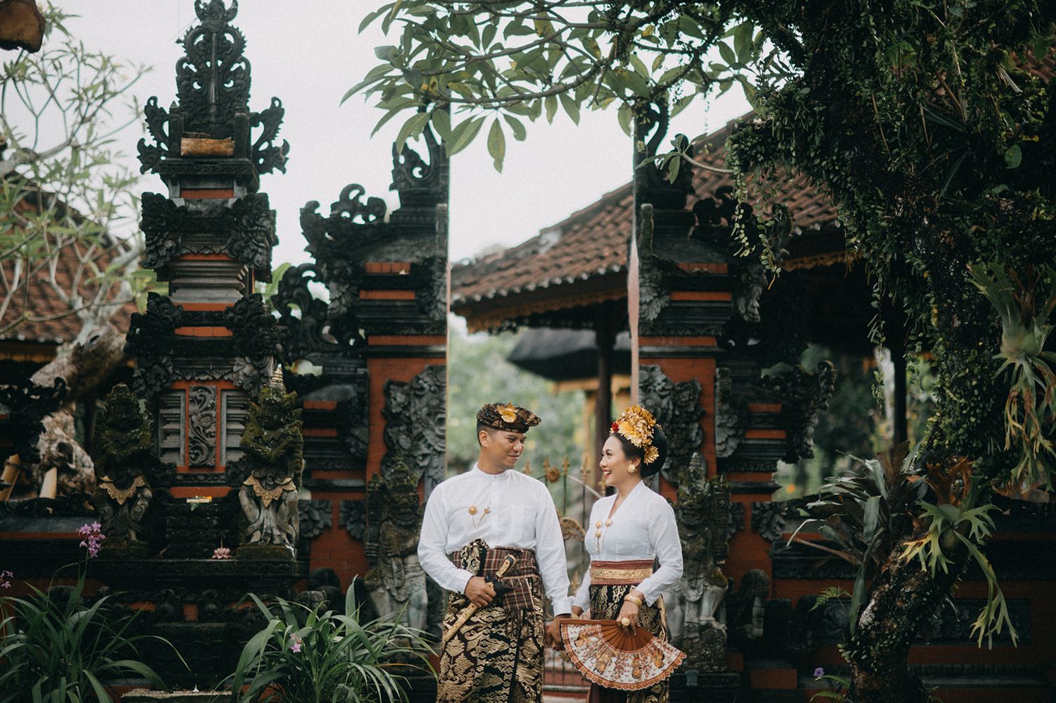 Couple in traditional Balinese attire posing at an ornate temple gate surrounded by lush tropical greenery.