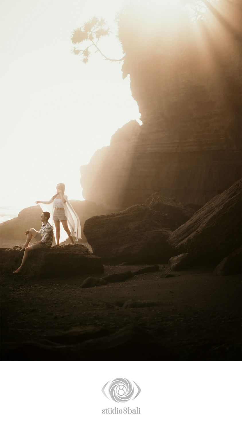Couple posing on rocky beach cliffs at golden hour with dramatic sunlight rays, captured by Studio 8 Bali.