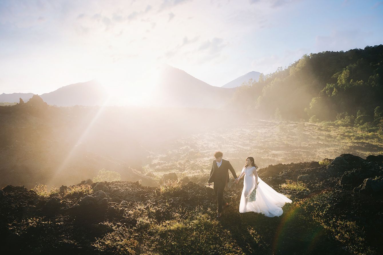 Bride and groom holding hands walking through a sunlit mountain landscape with dramatic golden backlight.