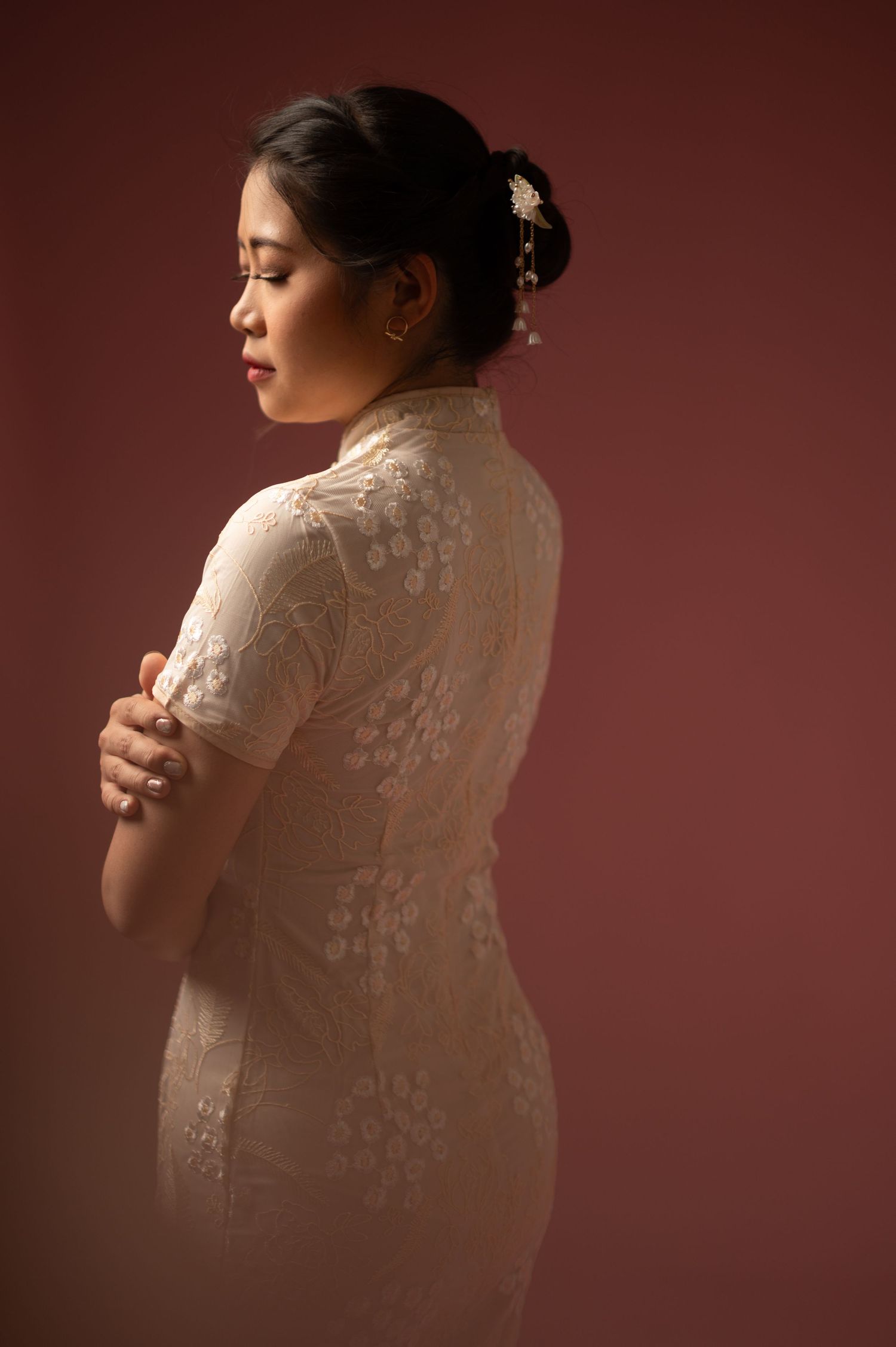 Woman in elegant white lace cheongsam dress with floral hair accessory, posed gracefully against a deep red background.