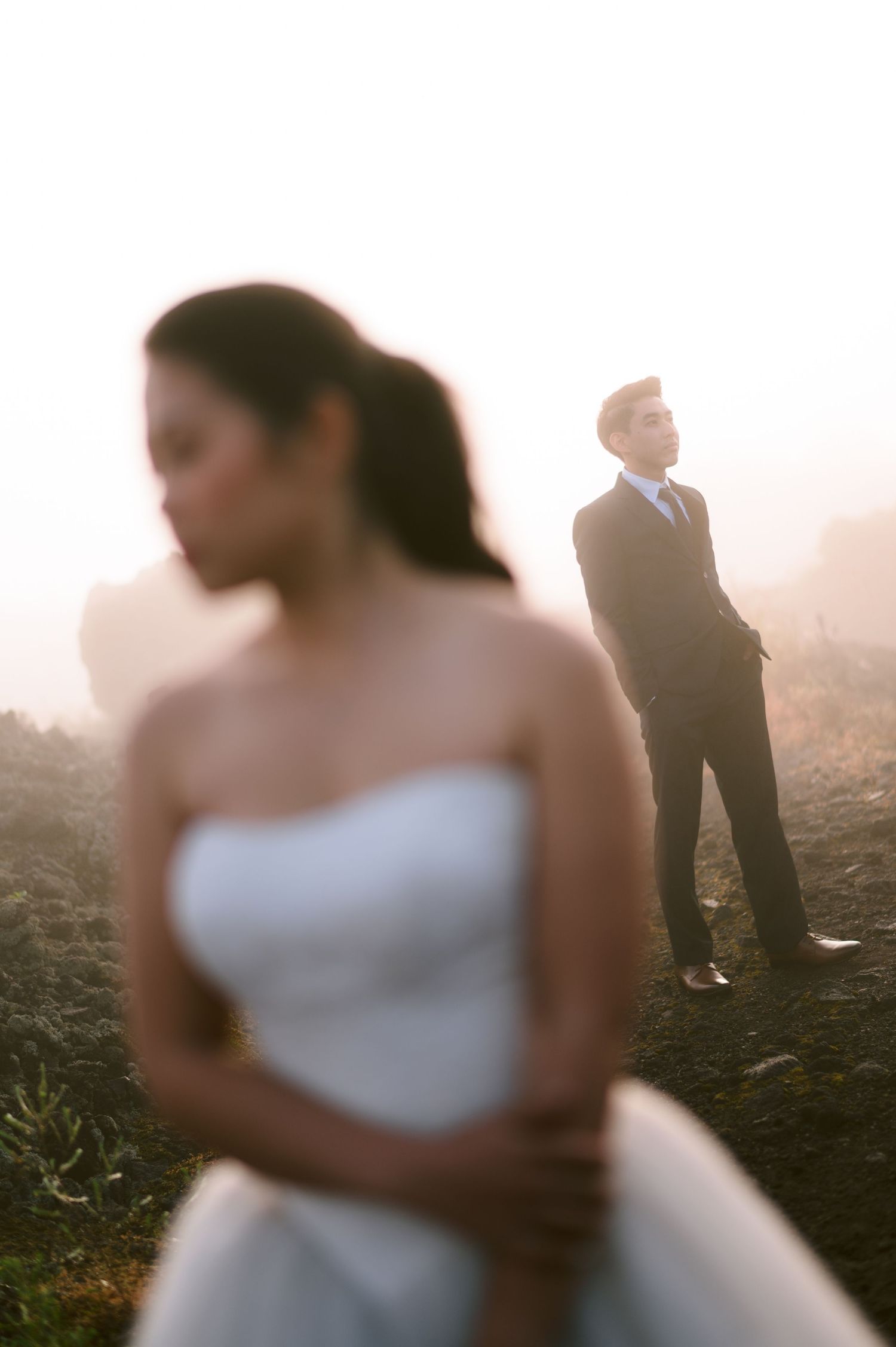 Bride in strapless white gown stands in foreground as groom in dark suit gazes upward in misty golden light.
