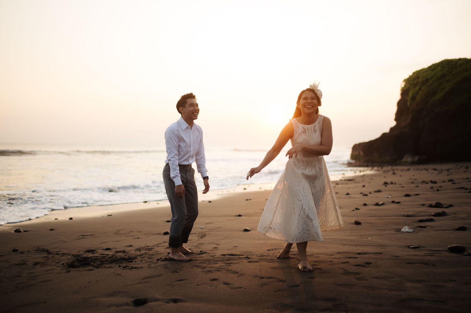 Couple holding hands walking on a sandy beach at sunset with ocean waves and rocky cliffs in the background.