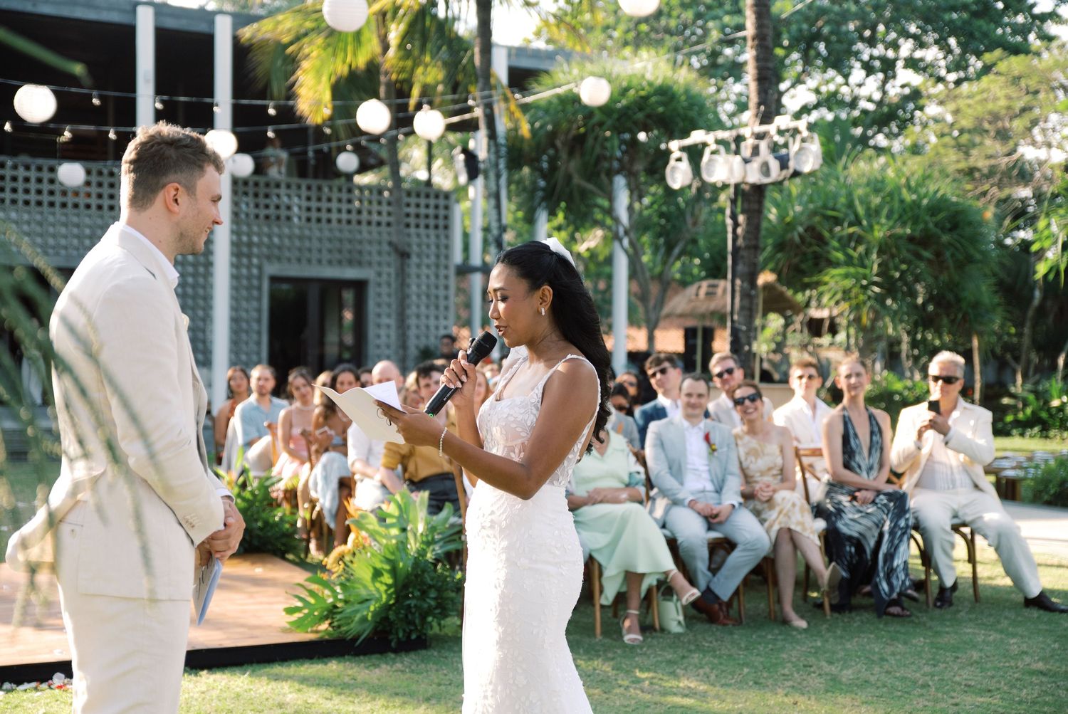 Bride speaks into microphone during outdoor tropical wedding ceremony while groom listens attentively.
