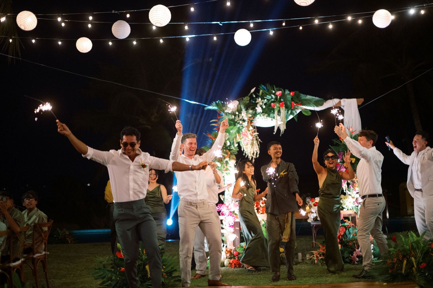 Groom and groomsmen celebrating with sparklers under string lights at an outdoor evening wedding reception.