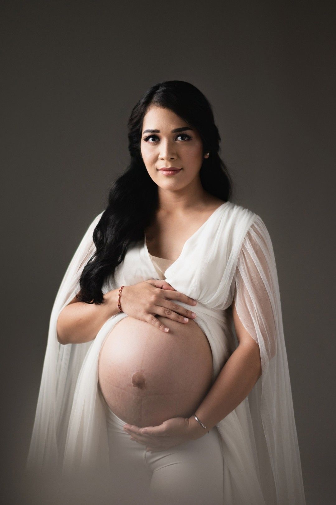 Pregnant woman in white sheer dress cradling her baby bump in a professional maternity studio portrait.