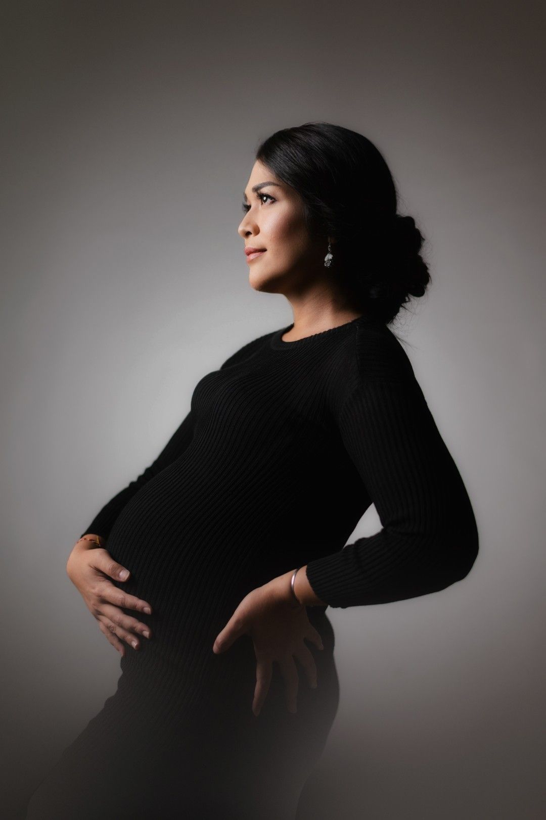 Pregnant woman in black dress cradling baby bump in elegant studio maternity portrait with gray background.
