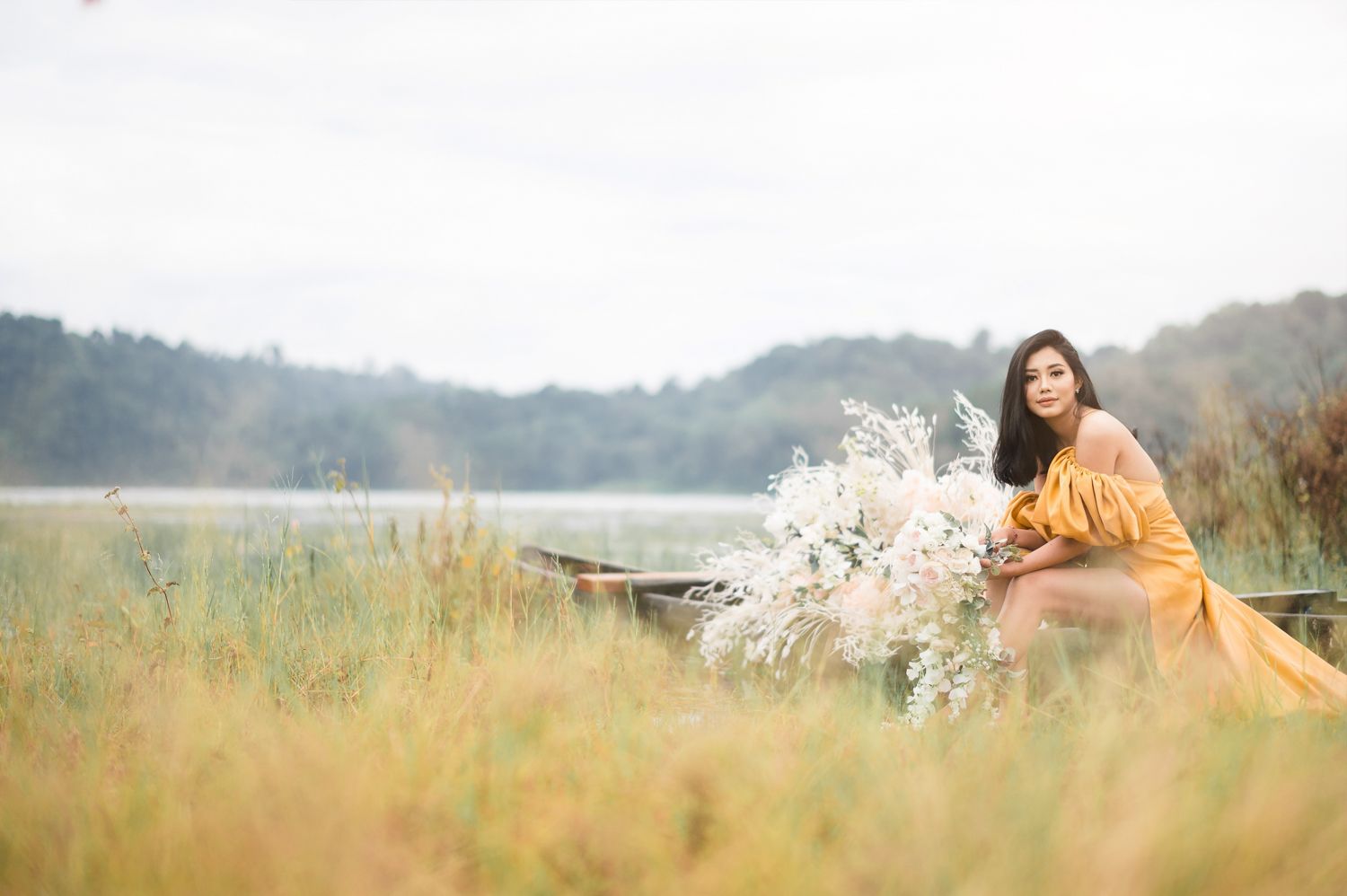Woman in yellow dress sitting in tall grass near a lake holding white wildflowers on a cloudy day.