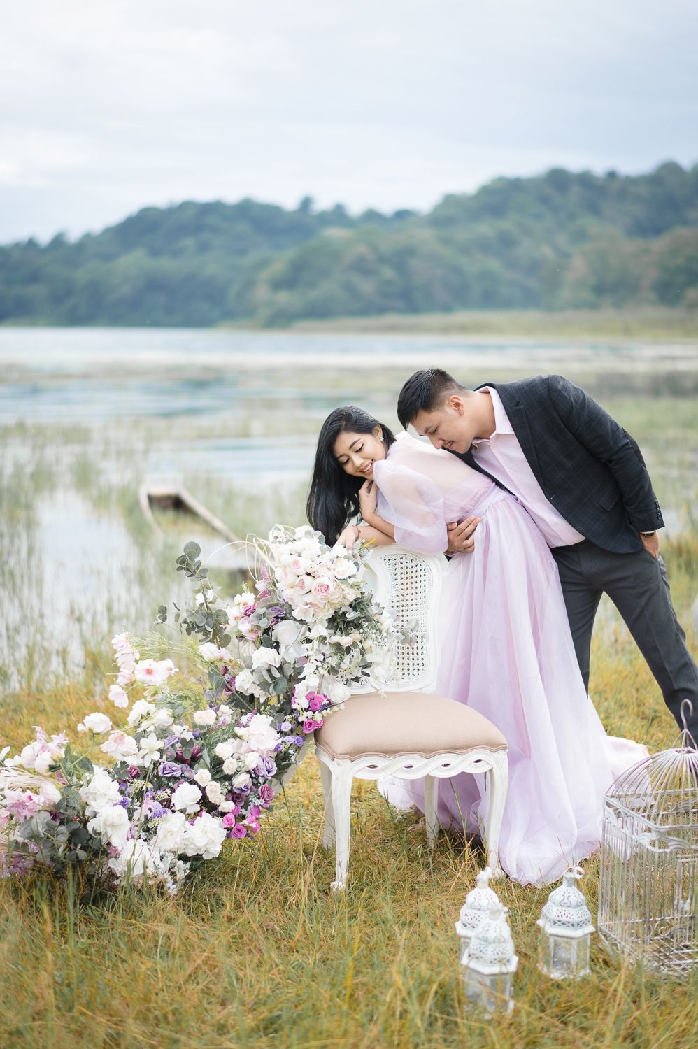 Couple in elegant attire embracing near a decorated chair with flowers by a serene lakeside.