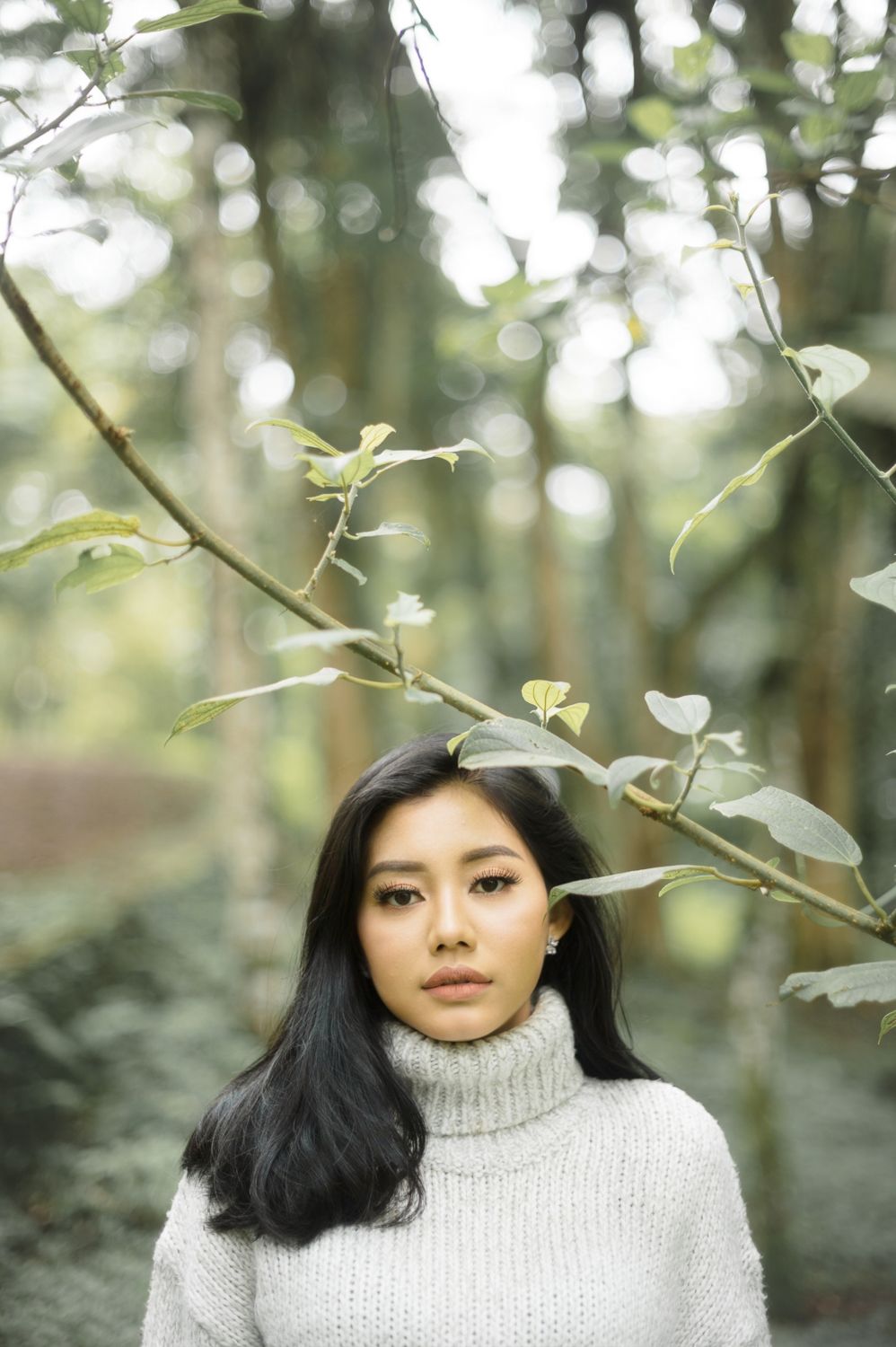Young Asian woman in gray turtleneck sweater standing among green forest branches, looking directly at camera.