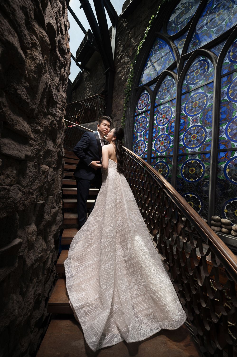 Couple in wedding attire sharing a romantic moment on ornate stairs beside stunning blue stained glass windows.