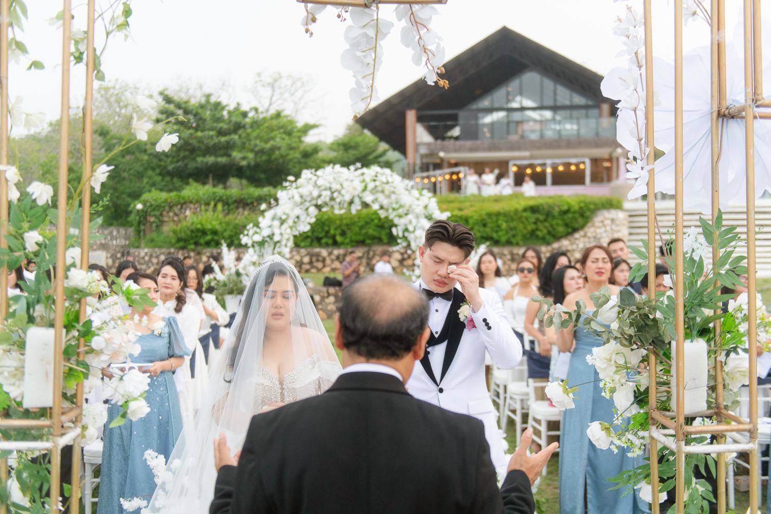 Outdoor wedding ceremony with couple at floral arch, guests in blue dresses, lush greenery and rustic venue backdrop.