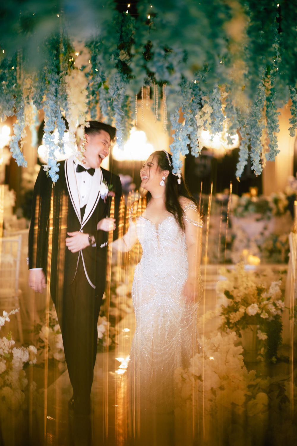 A joyful bride and groom laugh together under hanging floral arrangements at an elegant wedding reception.