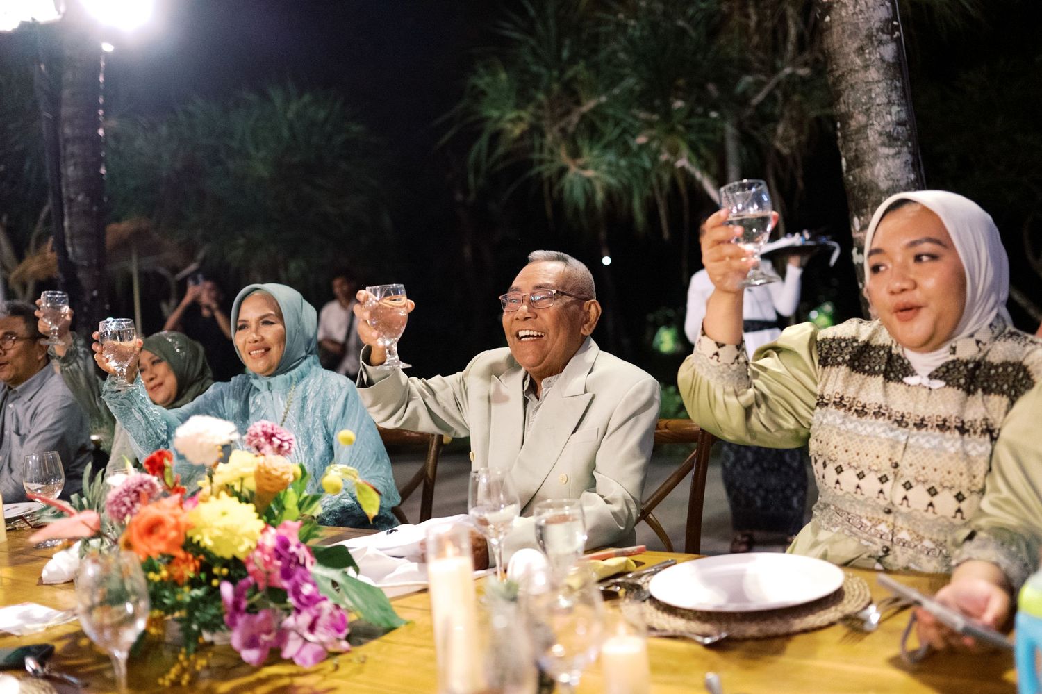 Wedding guests raising glasses in a toast at an elegant outdoor evening reception dinner.