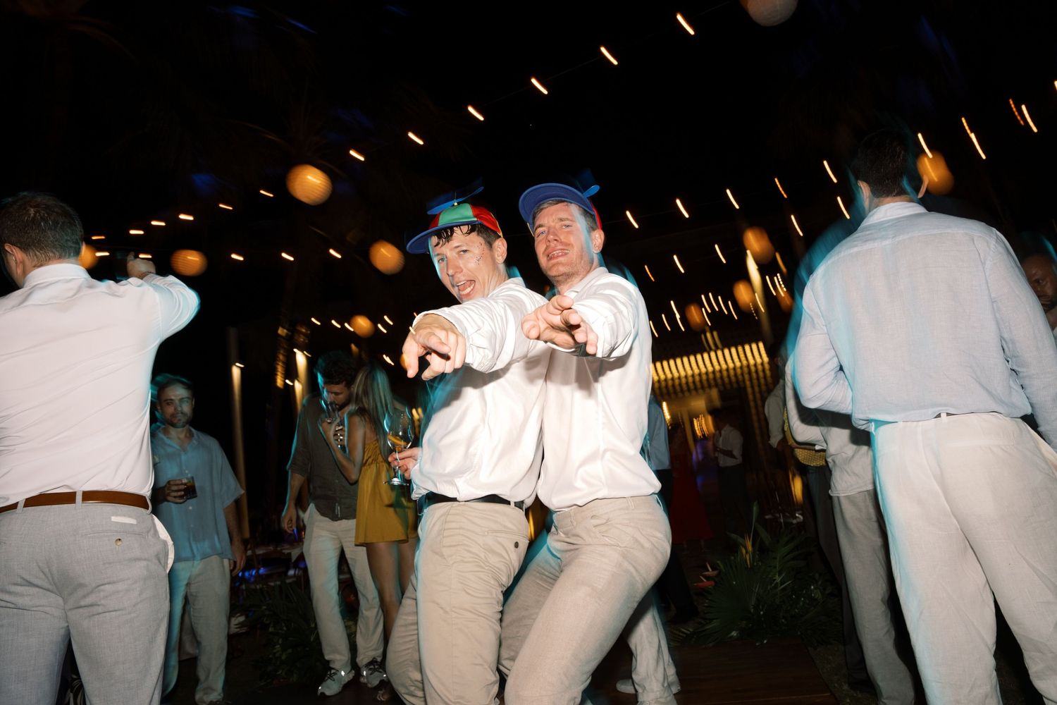 Two men in blue caps dance together at lively outdoor tropical wedding reception.