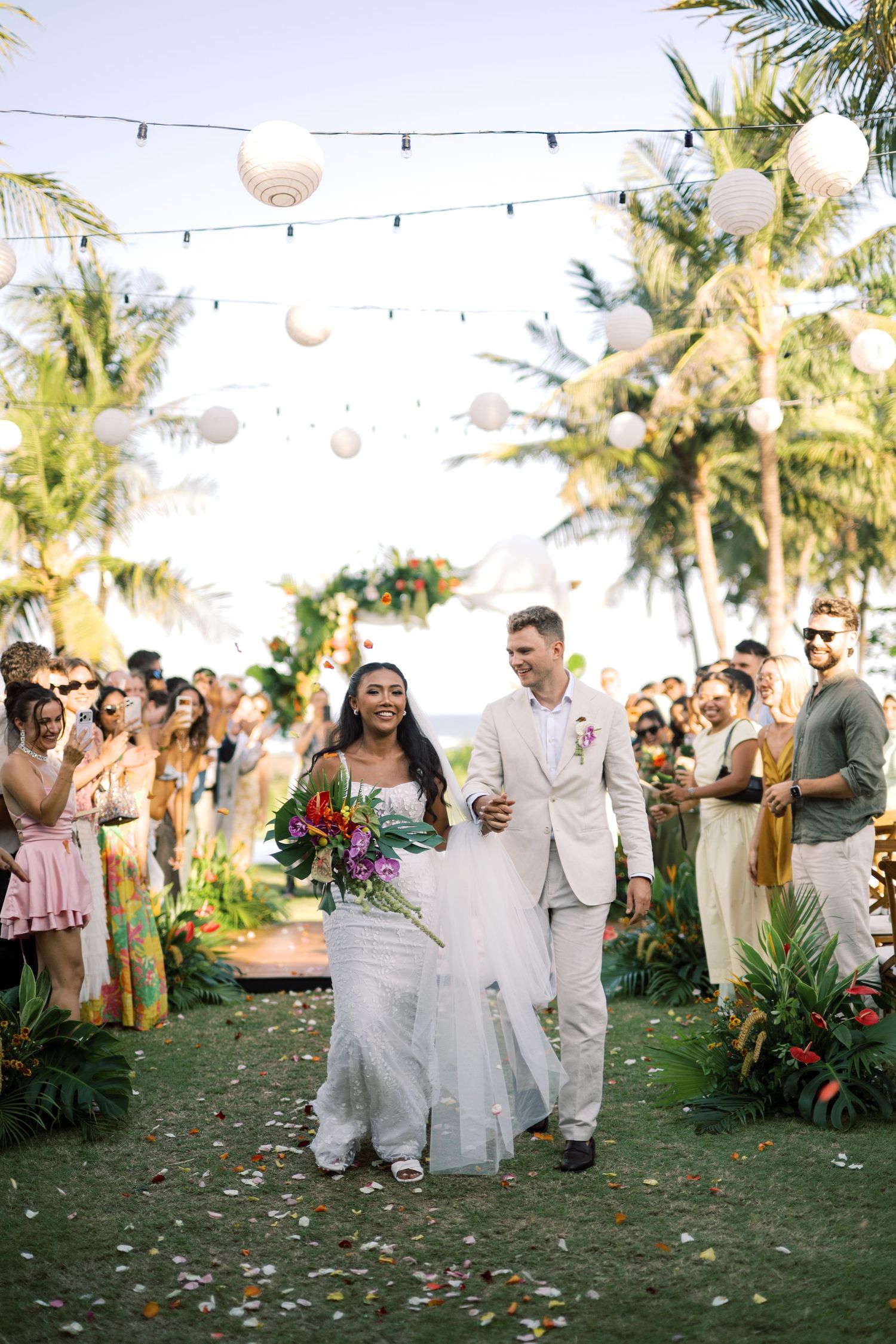 Bride and groom walk down aisle as guests cheer at tropical outdoor wedding reception.
