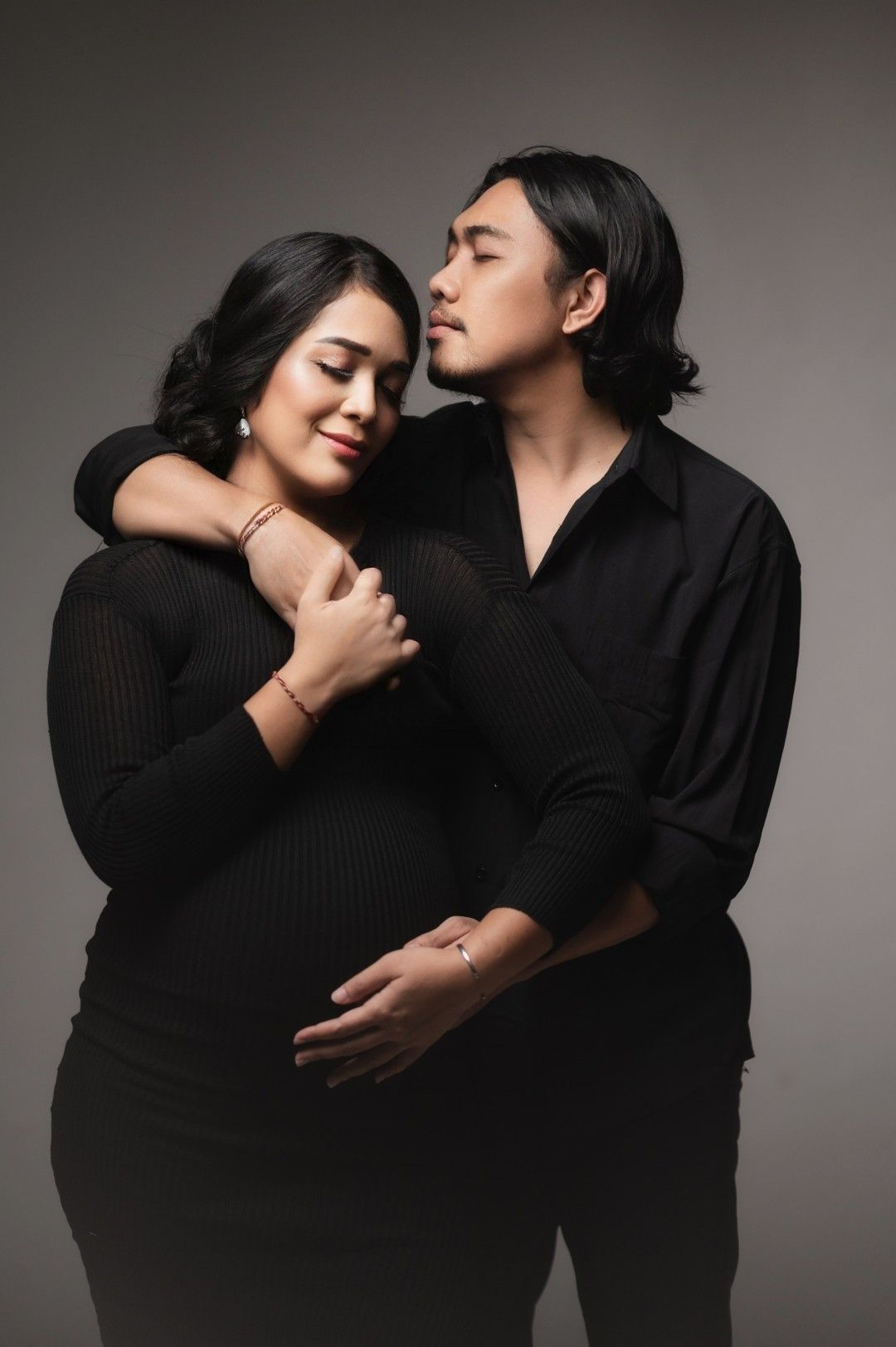 A couple in matching black outfits share a tender embrace, with the man gently holding the woman's pregnant belly.
