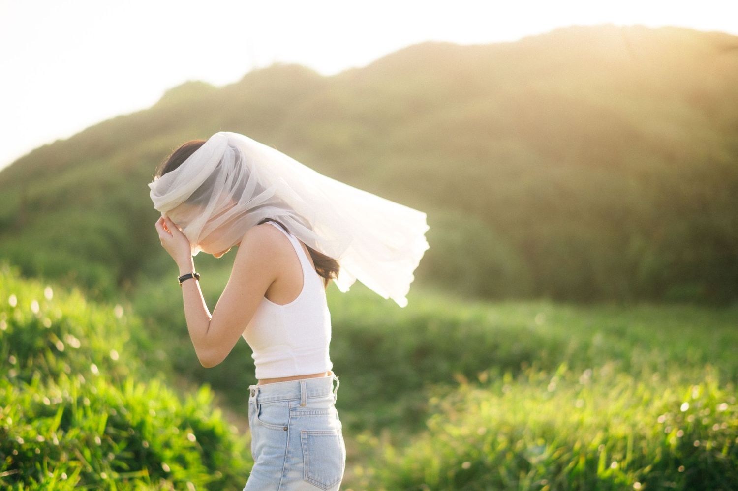 Woman in white top and jeans with a white veil blowing over her face in a sunny green landscape.