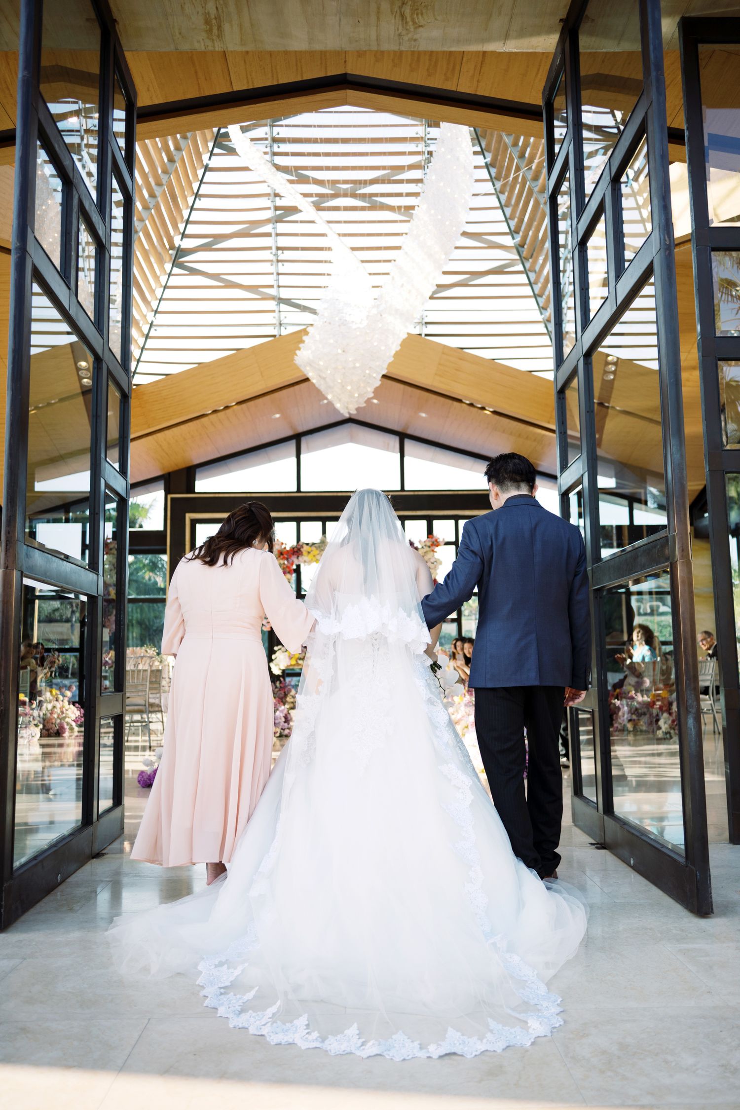 Bride in white gown and veil walks with family into elegant wedding venue with glass doors.