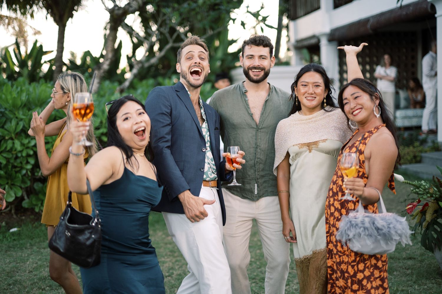 Group of friends smiling and raising glasses at an outdoor tropical party celebration.