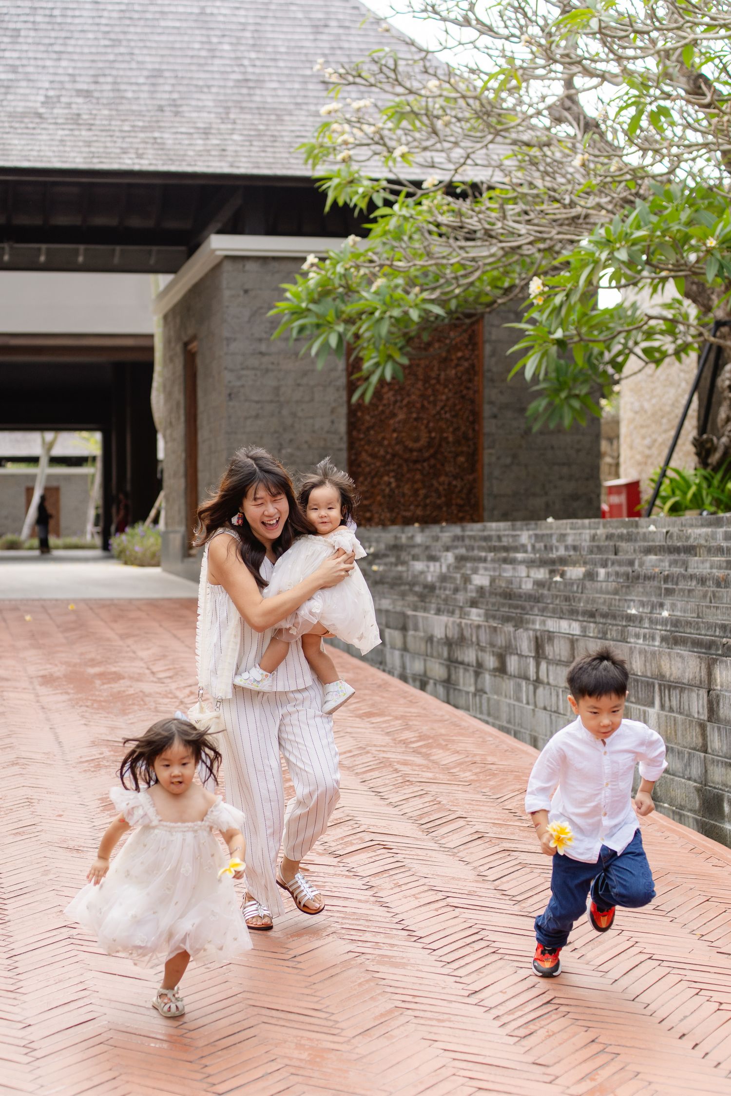 Asian mother holding baby while two young children run joyfully on brick pathway outside modern building.