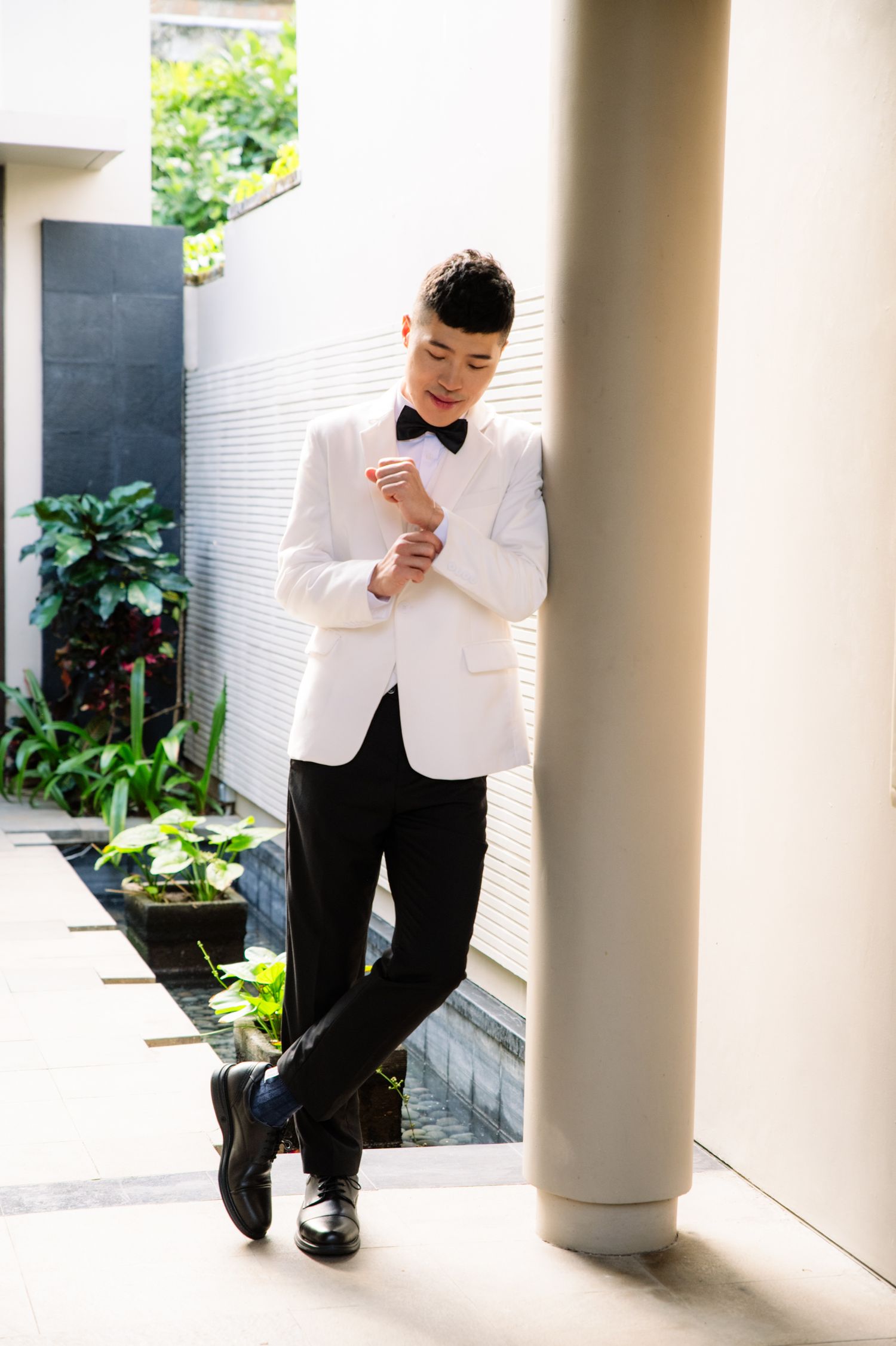 Man in a white tuxedo jacket, black bow tie and black trousers leans against a pillar while adjusting his cuff in a sunlit garden courtyard.