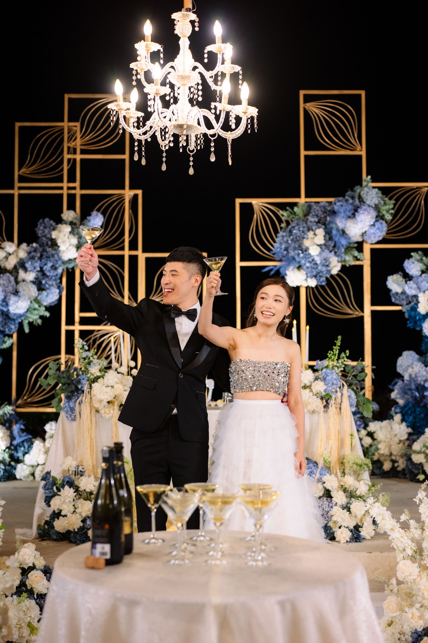 Bride and groom raise champagne glasses in a toast at a elegant wedding reception with a crystal chandelier and blue floral decor.