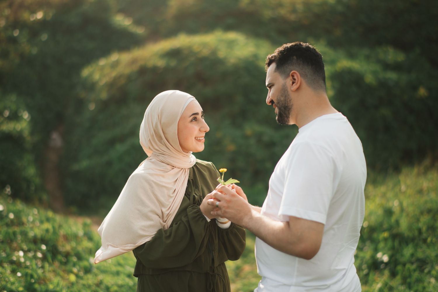 A couple in a green park, woman in hijab and man in white tee, hold hands and smile at each other.