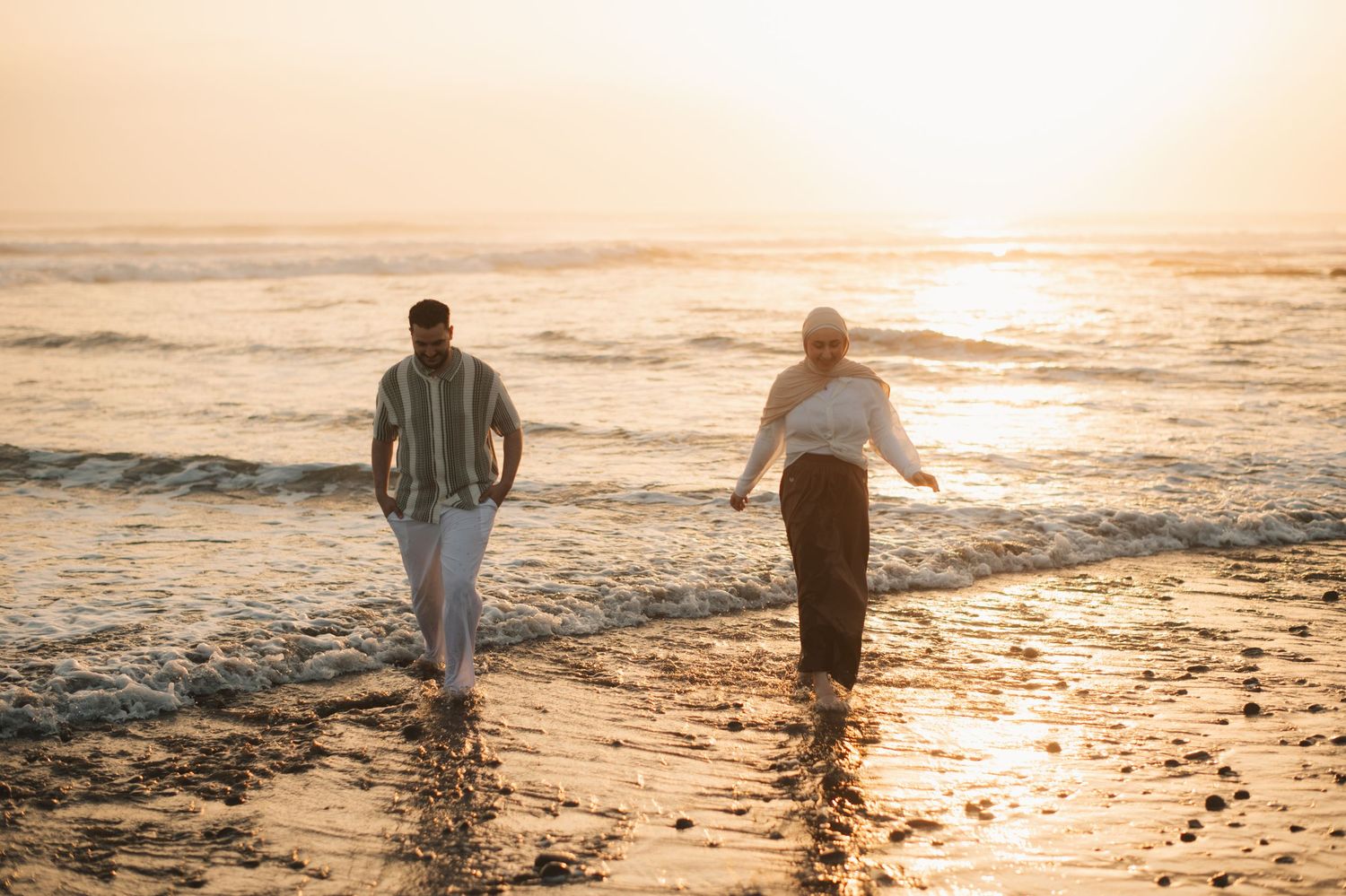 Two people walking barefoot along a sunlit beach at golden sunset, waves gently washing over the shore.