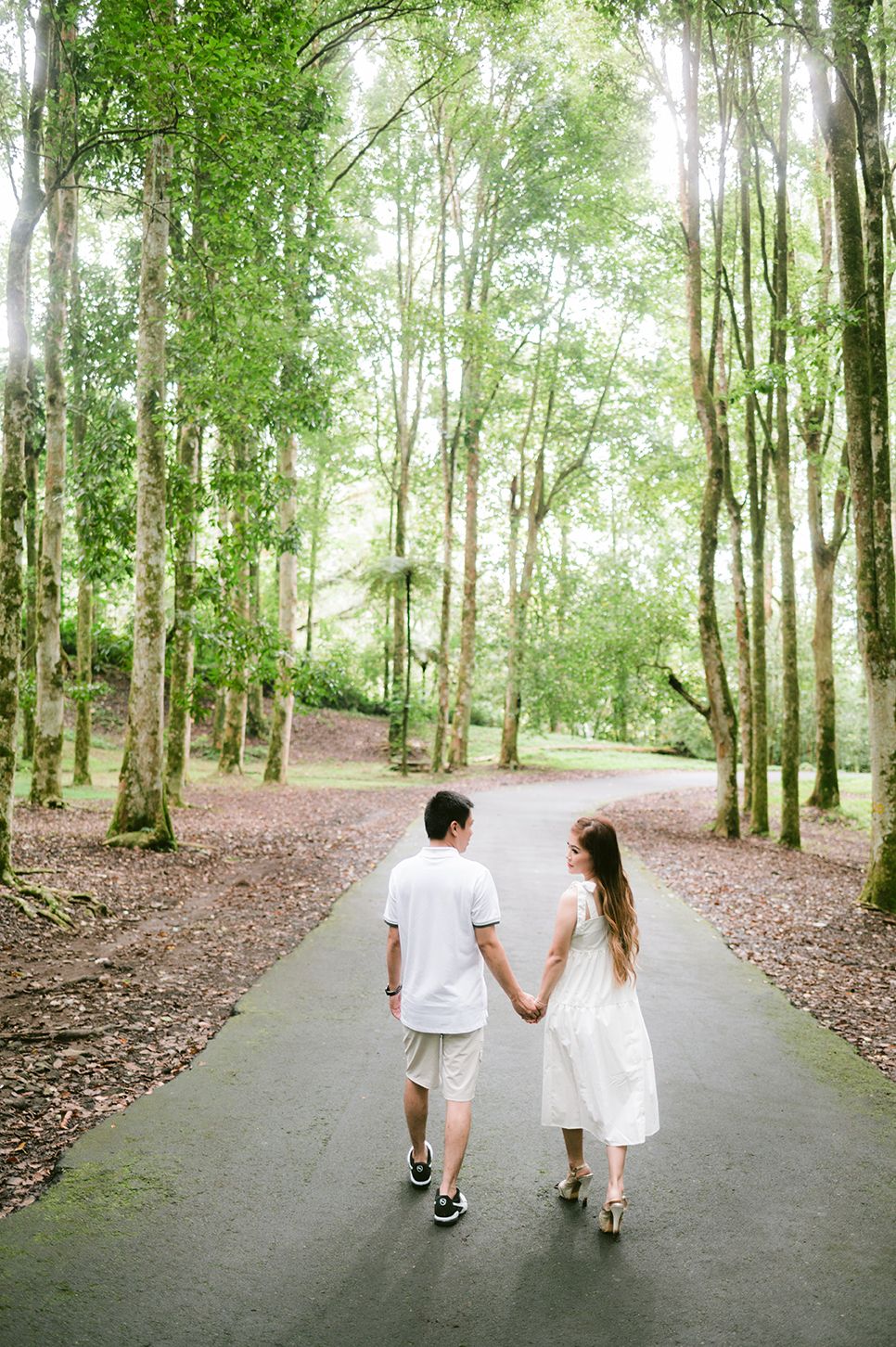Couple holding hands walking down a tree-lined path in a lush green forest, both dressed in white outfits.