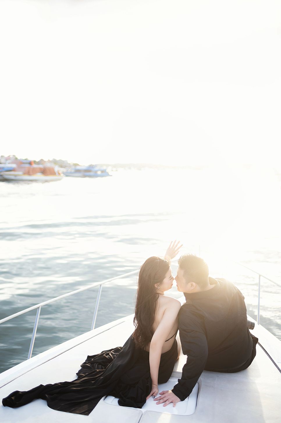 Couple in elegant black attire sharing a romantic moment on a yacht bow at sunset over calm waters.