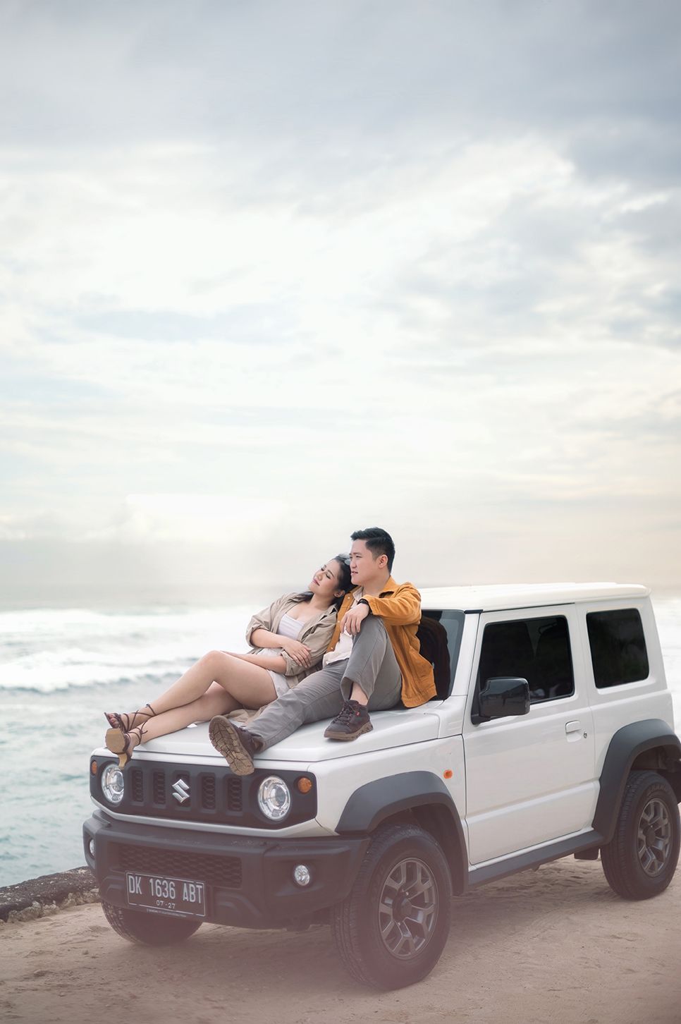 Couple sitting on hood of white Suzuki Jimny SUV on beach with ocean waves and cloudy sky backdrop.