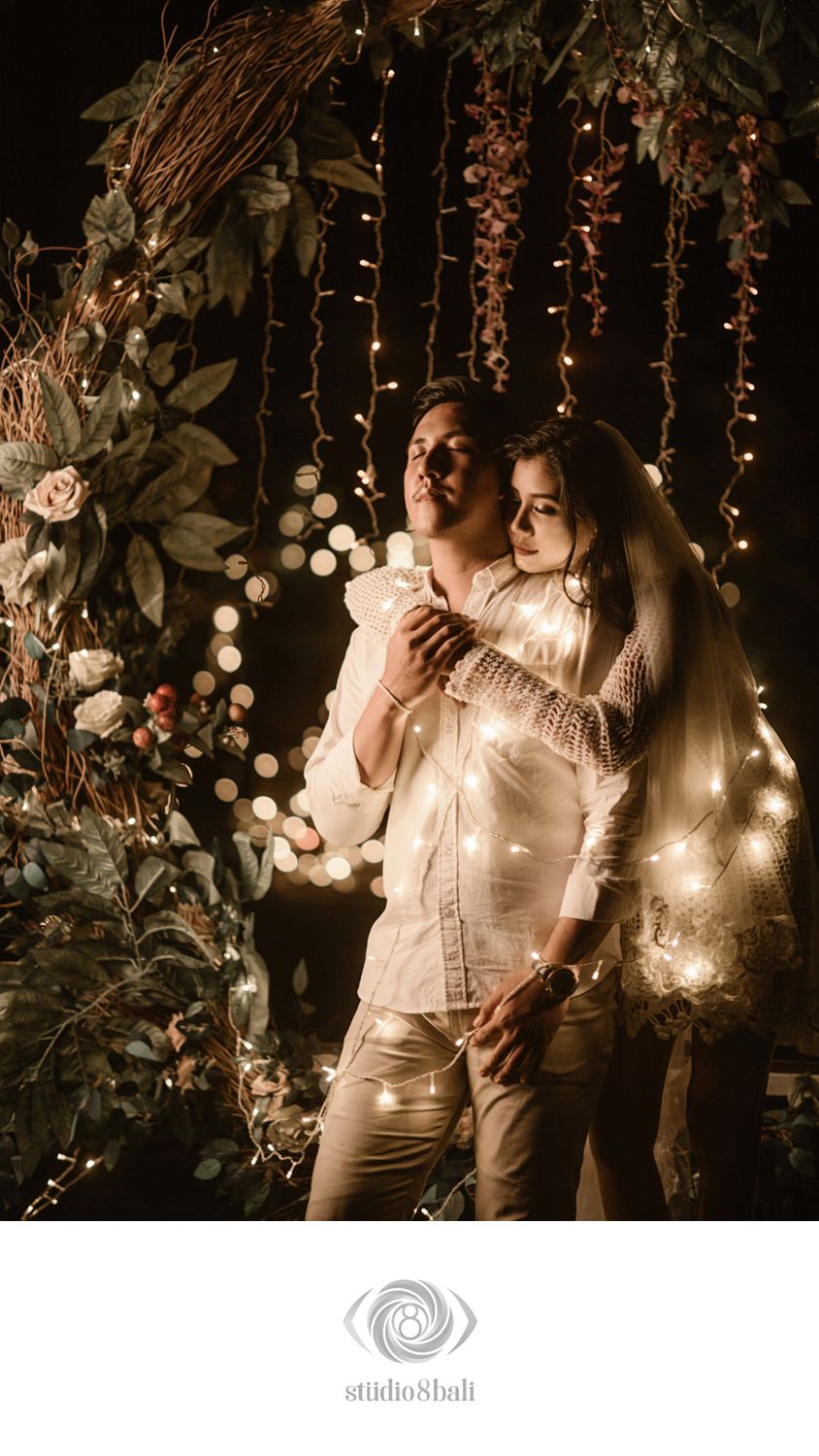 Romantic couple embracing under fairy lights and floral arch in a dreamy, bokeh-lit engagement photo by Studio 8 Bali.