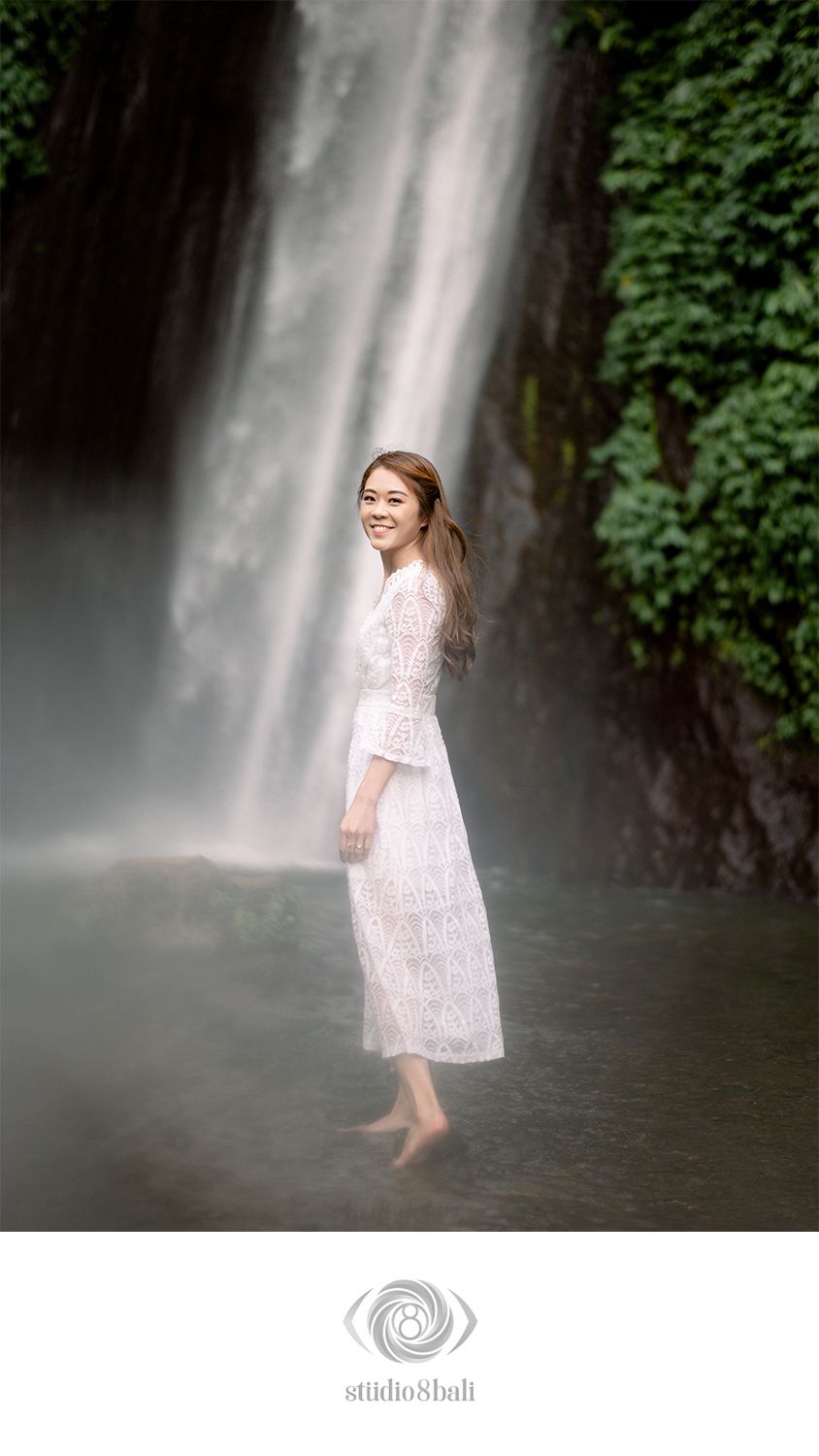 Smiling woman in white lace dress standing barefoot in misty waterfall pool surrounded by lush greenery in Bali.