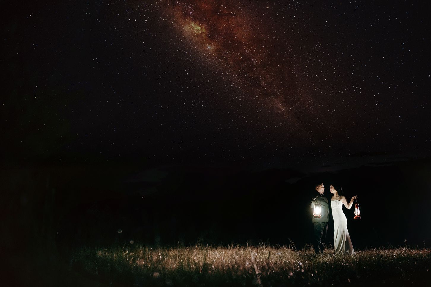 Couple holding a lantern in a field under a stunning Milky Way night sky.