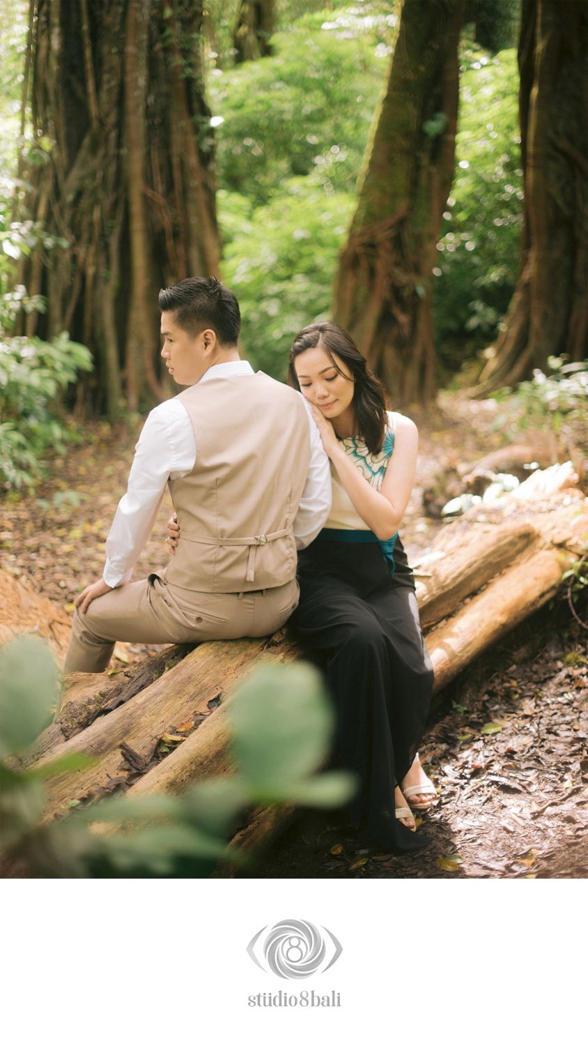 Couple sitting on a fallen log in a lush forest, woman resting her head on mans shoulder.