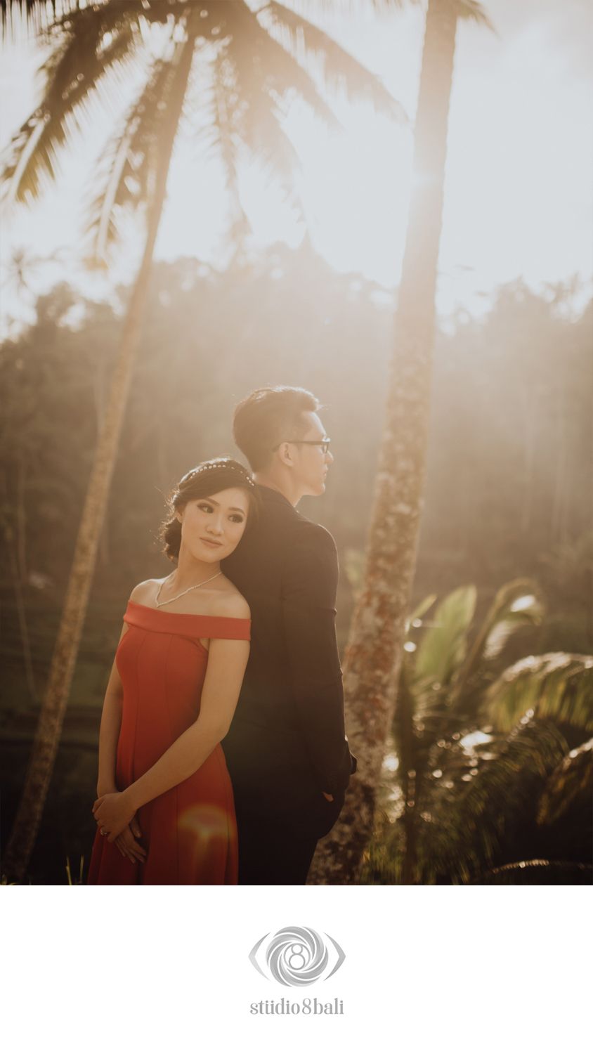 Couple poses romantically among tropical palm trees in golden sunset light, woman in red off-shoulder gown, man in black suit.
