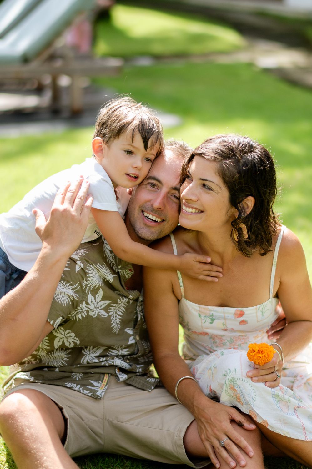 Happy family of three sitting on grass outdoors, father smiling with child hugging him and mother holding orange flower.