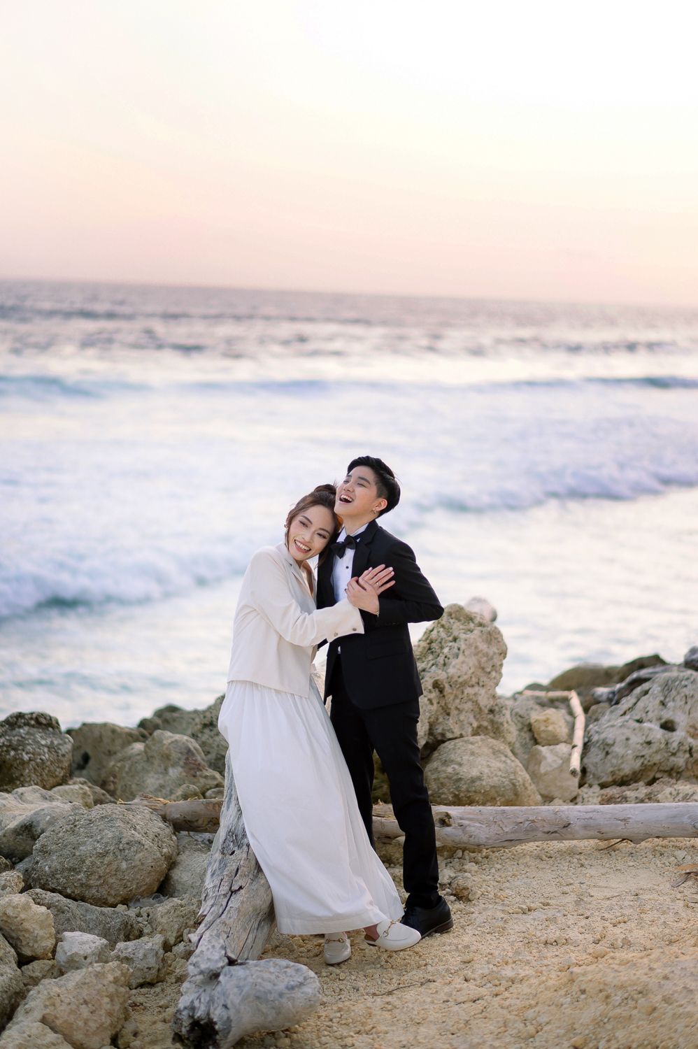 Couple in wedding attire embracing on rocky beach at sunset with ocean waves in background.