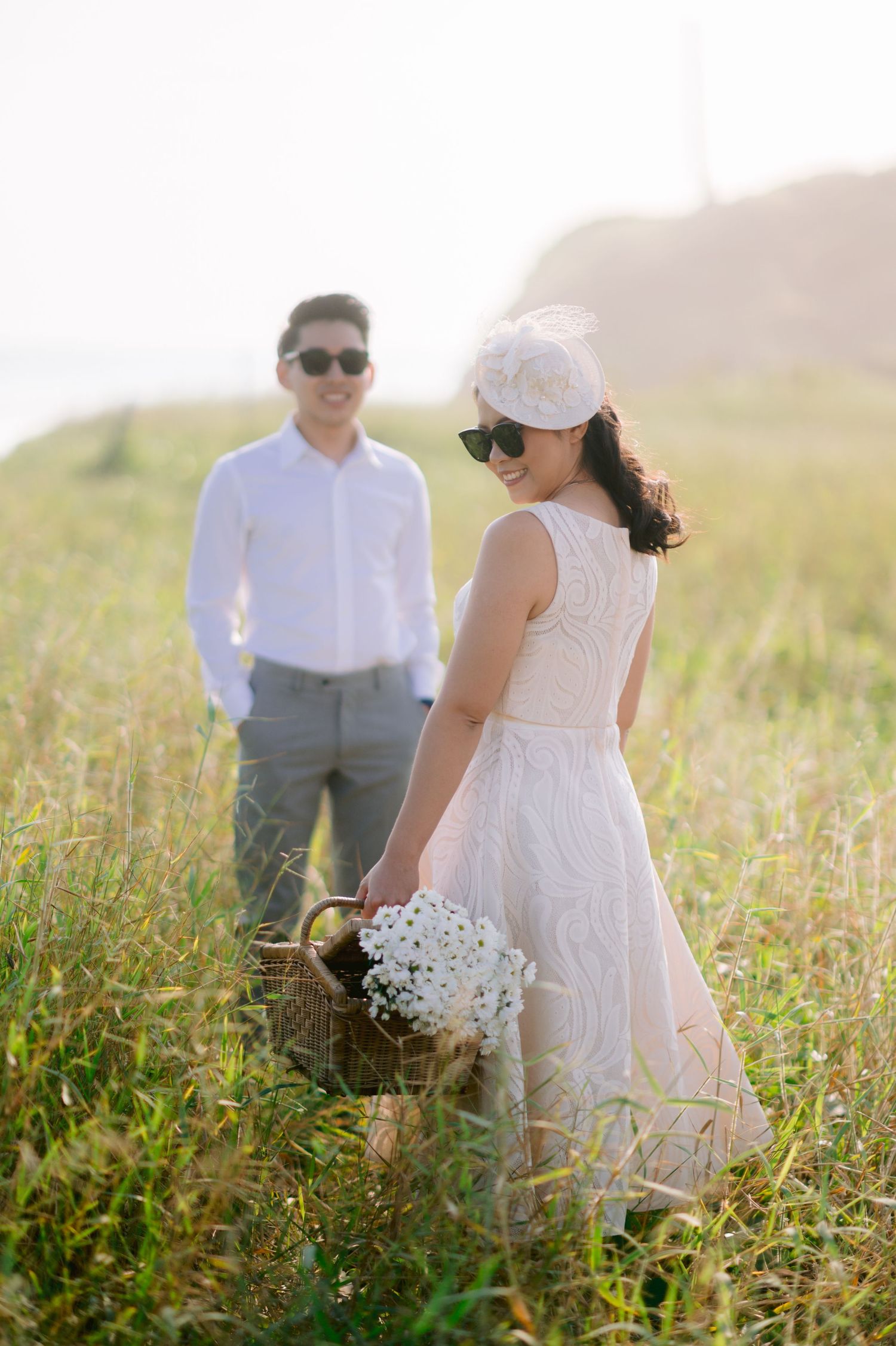 Couple in a sunny field, bride in white dress with fascinator holding flowers, groom in white shirt smiling in background.