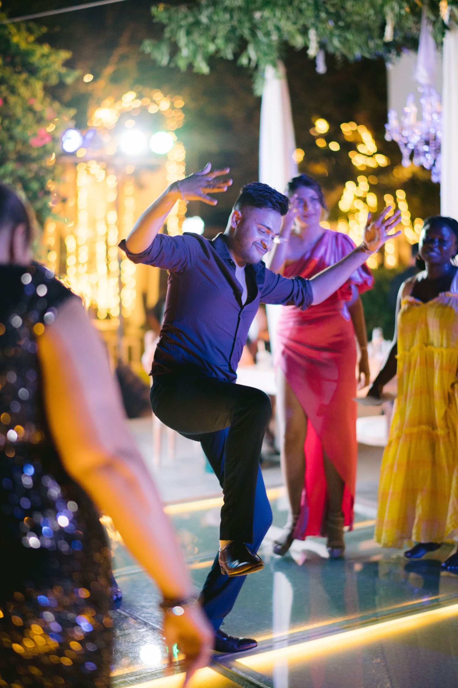 Man dancing energetically at an outdoor evening wedding reception with colorful lights and guests in festive attire.