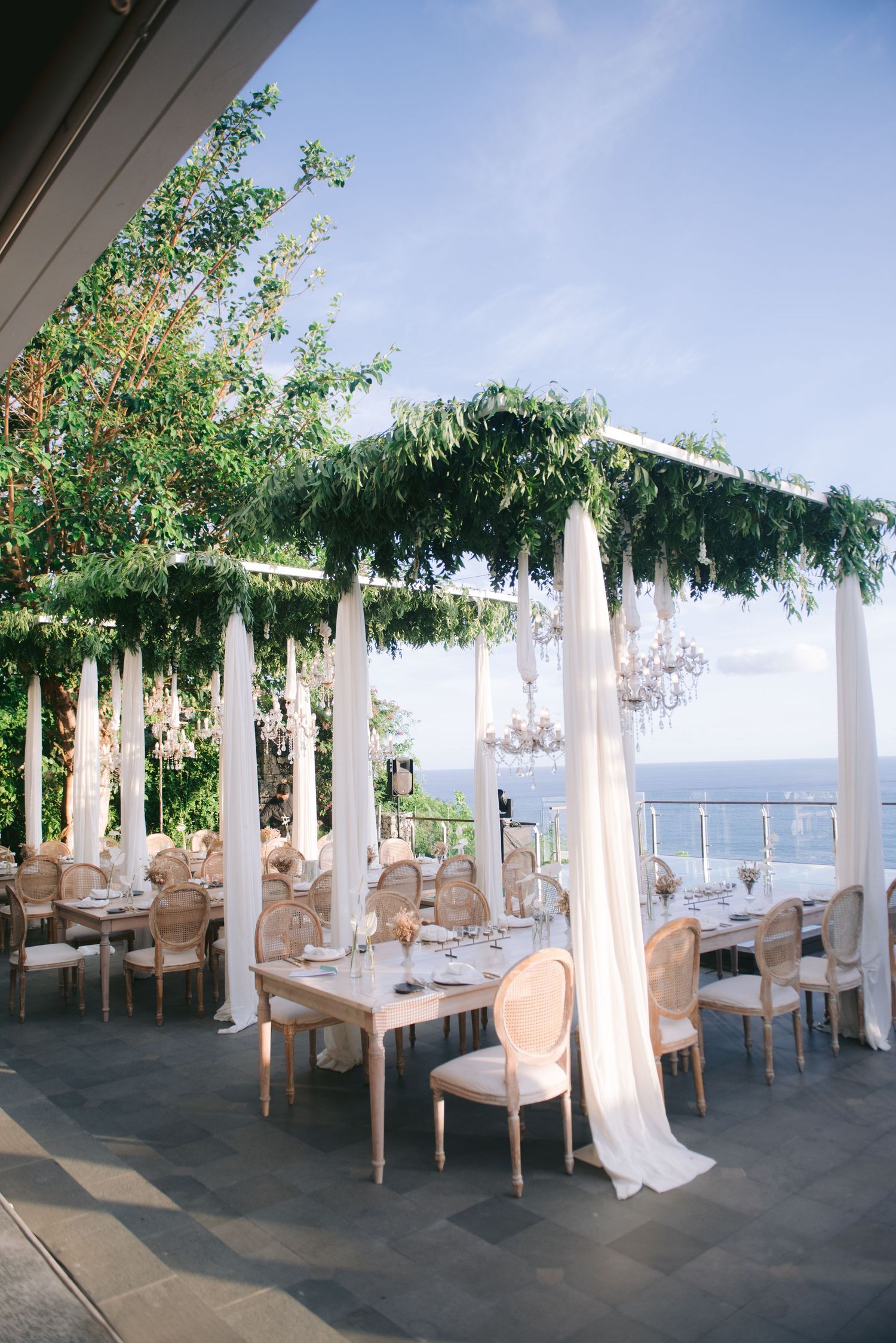 Elegant outdoor wedding reception with white draping, greenery canopy, chandeliers, and ocean views.