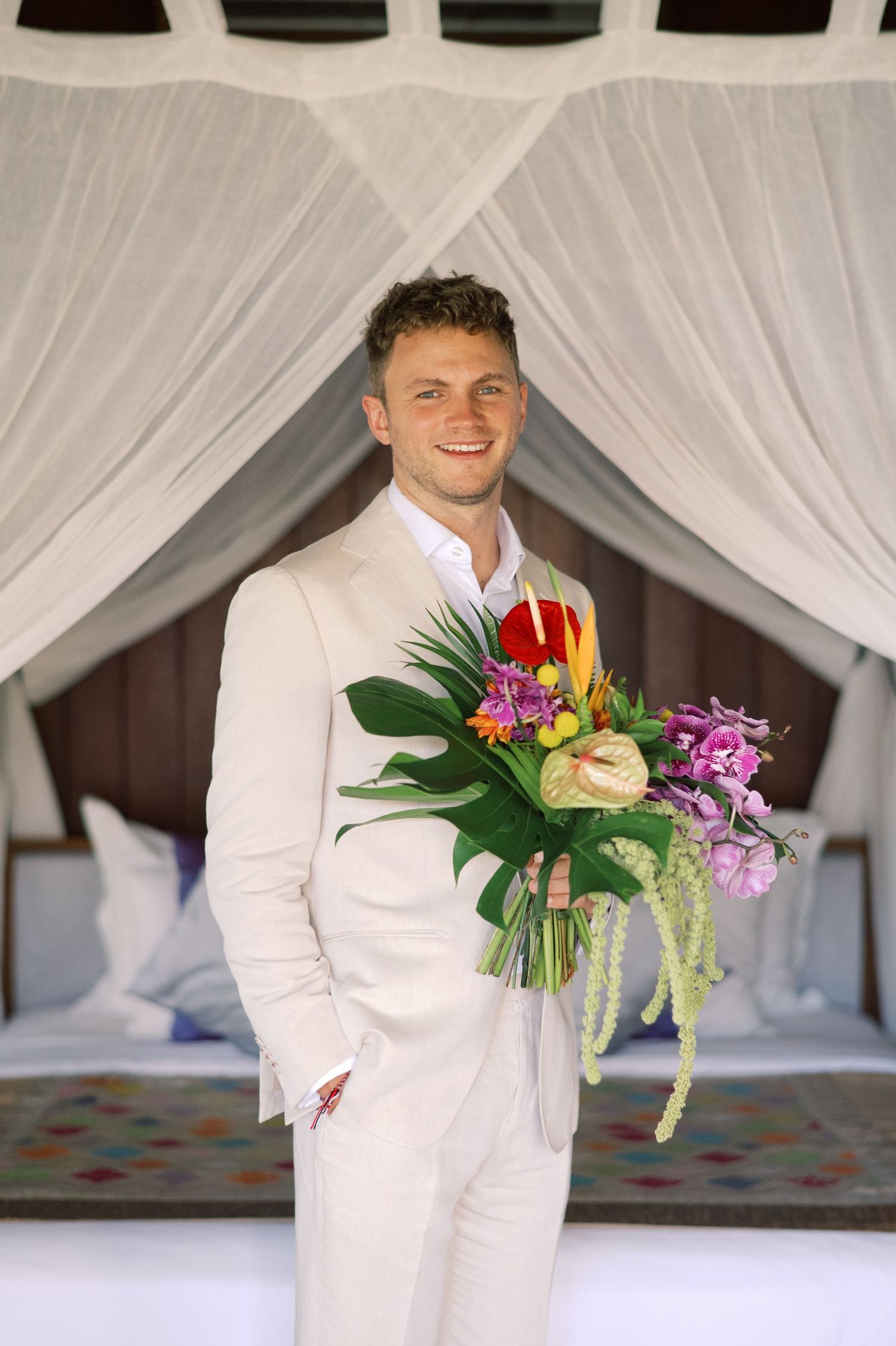 Smiling groom in white suit holding tropical wedding bouquet with colorful flowers under draped canopy.