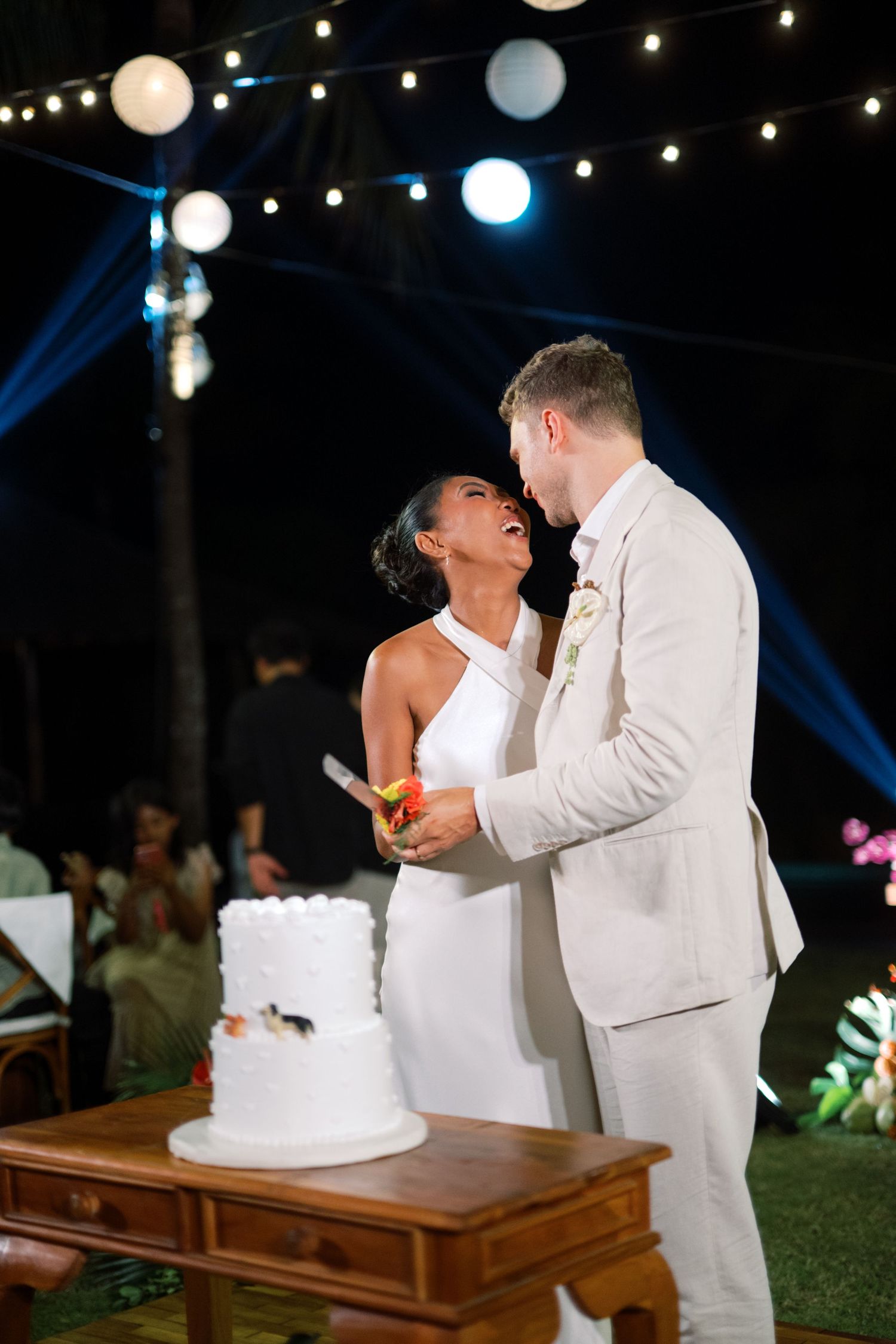 Bride and groom share a laugh while cutting white wedding cake under string lights at night.